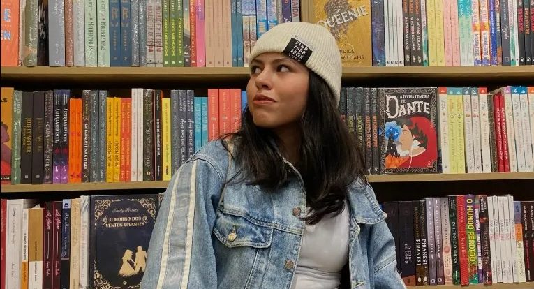 Foto colorida de Eduarda Costa. Ela é uma mulher negra de cabelos pretos e usa uma touca e um casaco jeans. Ao fundo, estante de livros