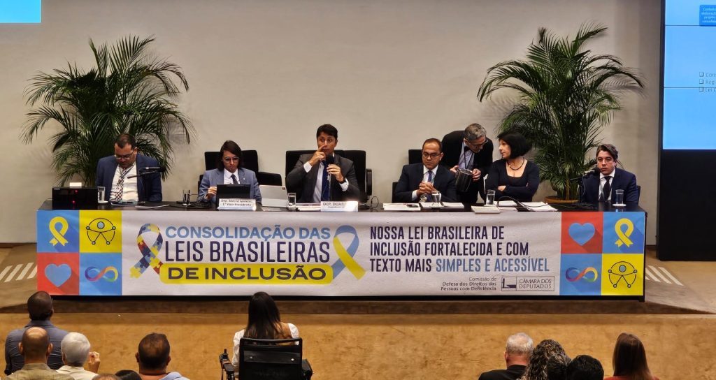 Foto colorida de audiência pública para discutir possível criação de código de consolidação das leis brasileiras de inclusão. Na imagem, aparece uma bancada com a presença do deputado Duarte Jr no centro e com o microfone