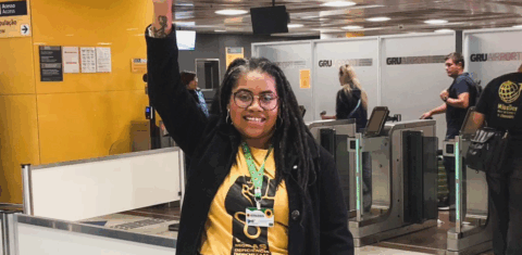 Foto colorida de Luciana Viegas. Luciana é uma mulher preta, com cabelos trançados. Ela sorri e levanta o braço direito. Ela usa um cordão de girassol, uma camiseta amarelo e um casaco preto. Ao fundo, aeroporto.