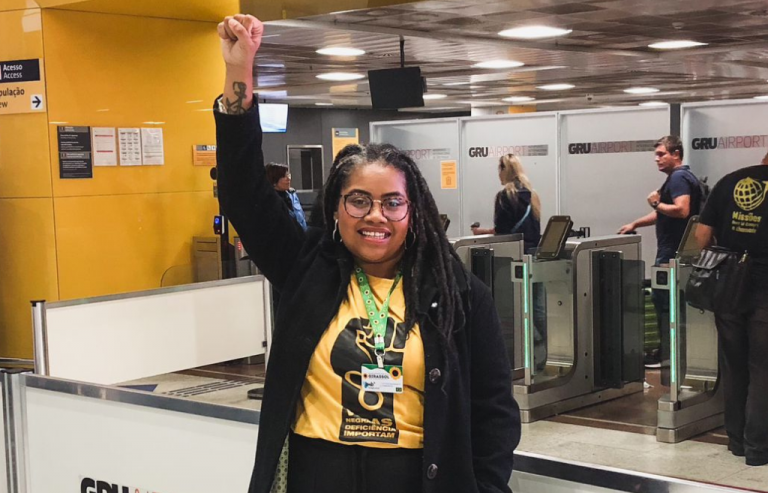 Foto colorida de Luciana Viegas. Luciana é uma mulher preta, com cabelos trançados. Ela sorri e levanta o braço direito. Ela usa um cordão de girassol, uma camiseta amarelo e um casaco preto. Ao fundo, aeroporto.