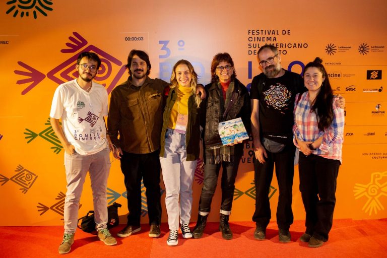 Foto colorida de equipe do "Sobre a cabeça, os aviões" no 3° Bonito Cinesur, onde o filme foi premiado. Eles aparecem em pé, posando para a foto em frente ao banner do festival