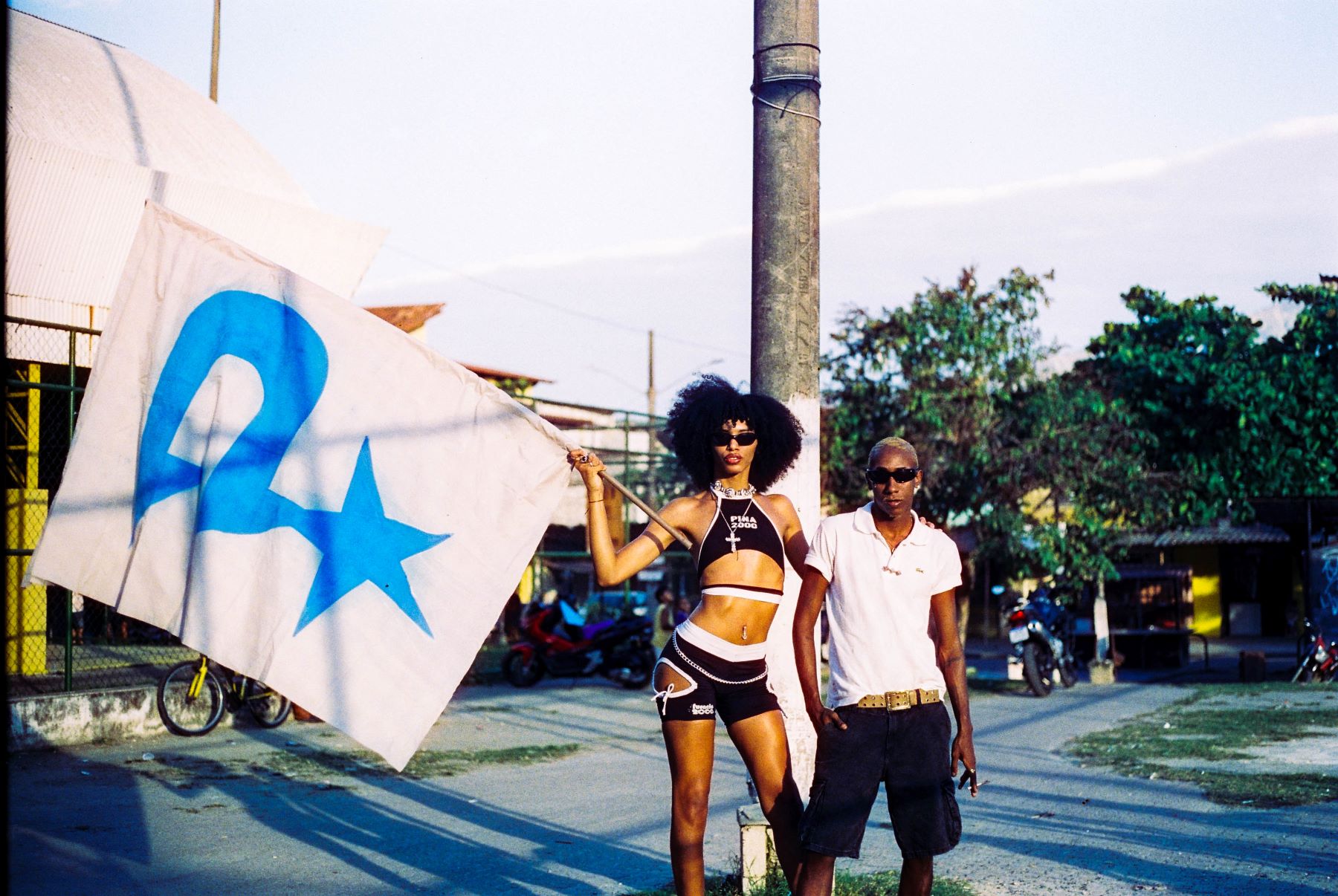 O estilista Abacaxi, na Vila Kennedy, ao lado da modelo Yasmin Boni, com uma bandeira com a logo da Piña: As marcas querem o que a favela produz, mas não querem o favelado" (Foto: Luiza Trindade / Divulgação)