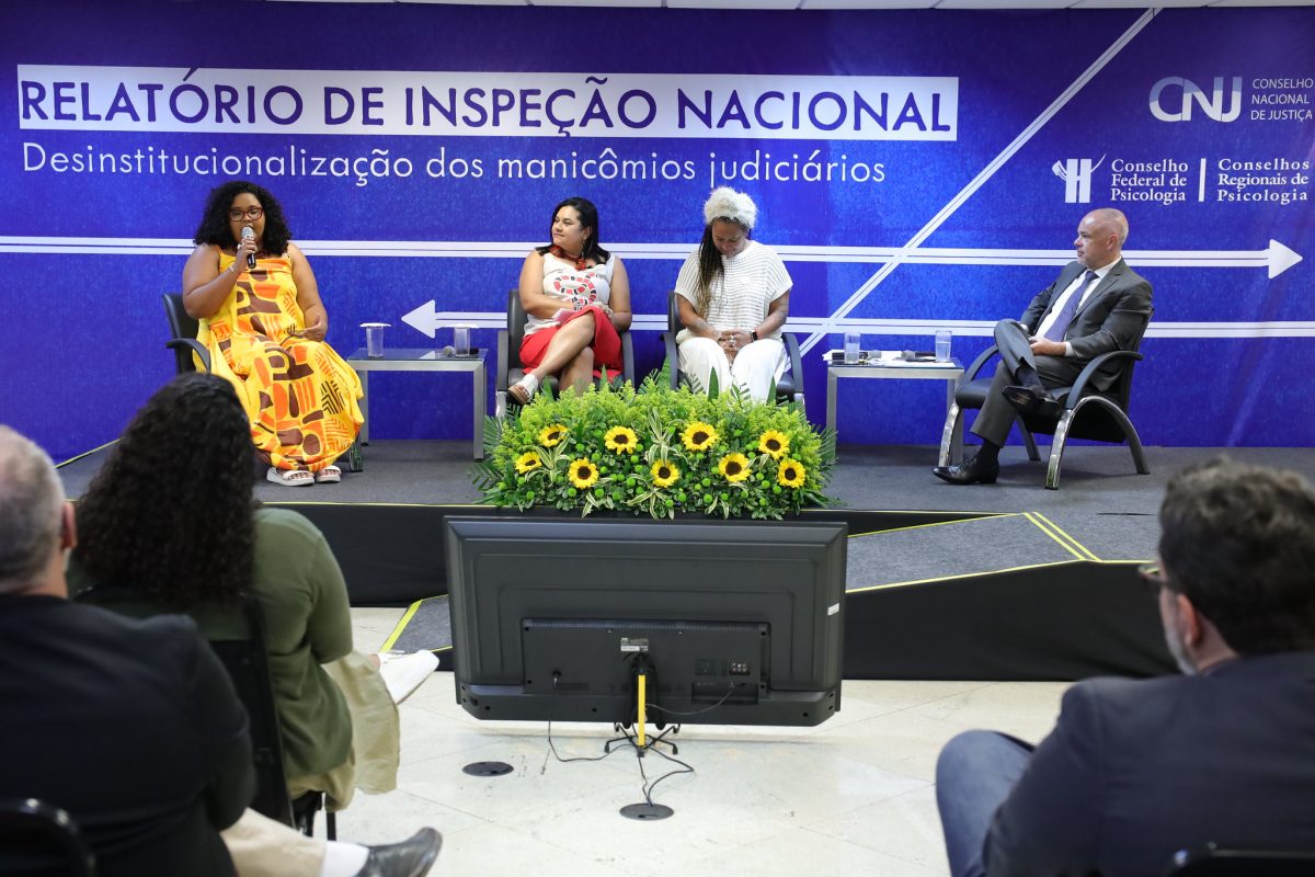 Foto colorida de lançamento do relatório sobre manicômios judiciários do CFP. Na imagem, aparecem quatro pessoas, três mulheres e um homem no palco. Uma delas, à esquerda, está com o microfone. Ao fundo, parede com a frase Relatório de Inspeção Nacional. Na frente do palco, aparece uma série de girassois.