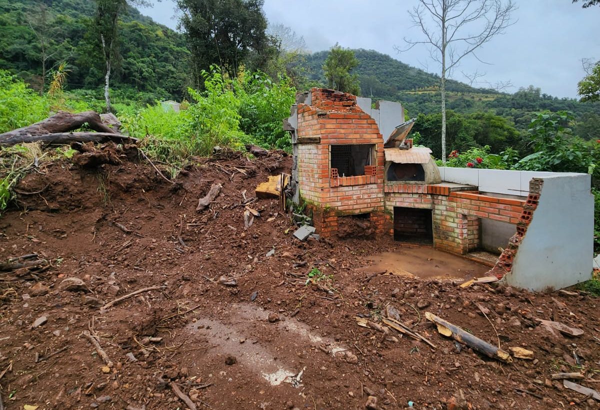 Foto colorida de casa atingida por deslizamento em Santa Tereza. Na imagem, aparece vestígios de uma churrasqueira em meio a muita terra. Ao fundo, diversos morros