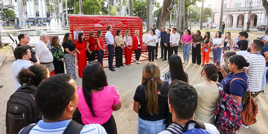 Foto colorida de inauguração de um banco vermelho gigante em Caruaru (PE). Na imagem, diversas pessoas aparecem reunidas em torno do banco