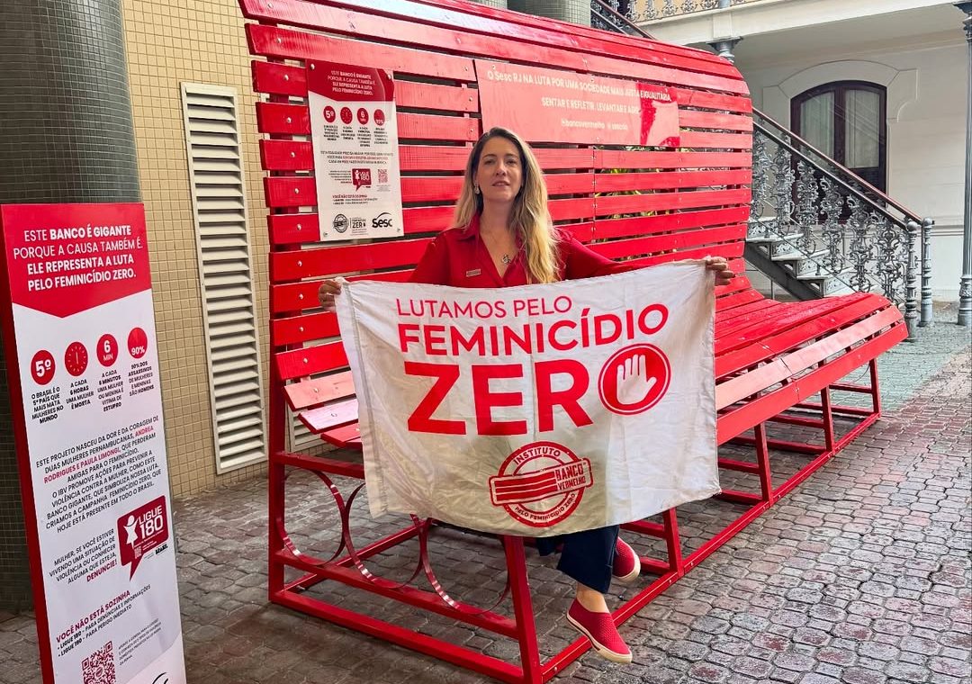 Foto colorida de Paula Limongi sentada em um banco vermelho gigante. Ela é uma mulher branca, loira e está segurando um cartaz que diz "Feminicídio zero", com a logo do Instituto Banco Vermelho