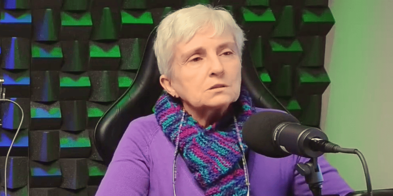 Foto colorida de Dolores Sala. Ela é uma mulher branca com cabelos brancos curtos. Ela usa um cachecol azul com roxo e uma blusa rocha e em sua frente aparece um microfone