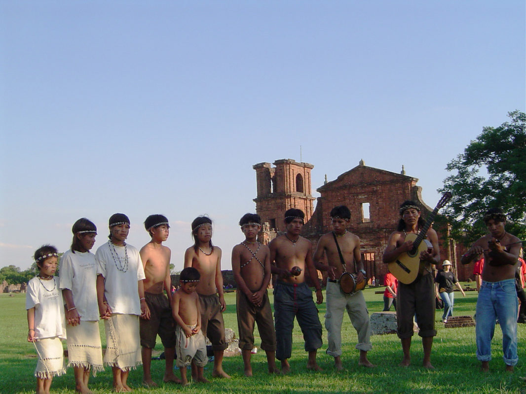 Foto colorida de indígenas guaranis no Sítio Arqueológico de São Miguel das Missões. Na imagem, os guaranis aparecem reunidos e muitos sem camisa. Ao fundo, aparecem as ruínas das Missões