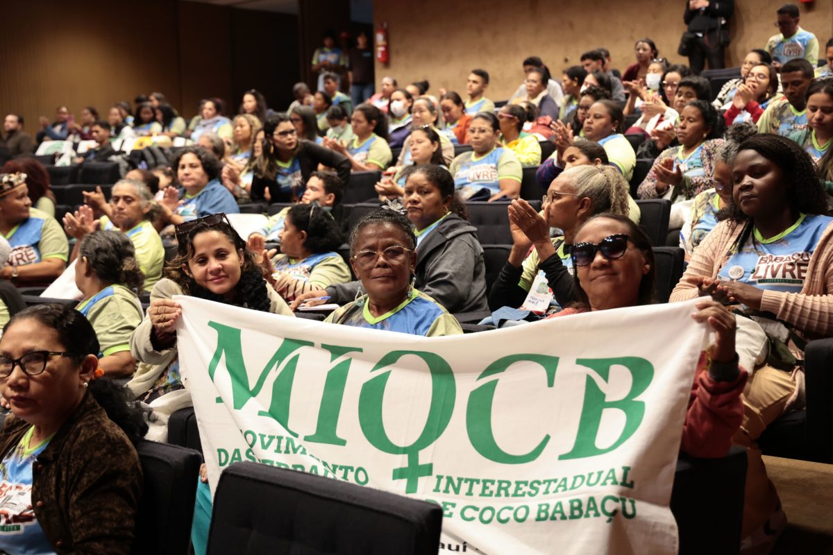 Foto colorida de três mulheres segurando uma bandeira do MIQCB, com a sigla do movimento escrita em verde sob fundo branco. Ao fundo da imagem, diversas pessoas em auditório
