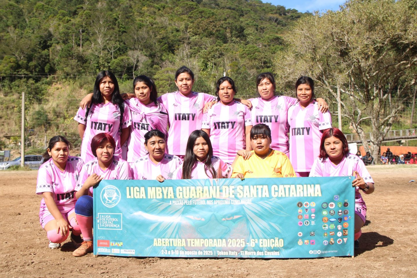 Foto colorida das atletas da Kunhangue Itaty reunidas para foto oficial antes de partida. Elam usam uniforme rosa e seguram uma faixa da Liga Mbya Guarani de Santa Catarina