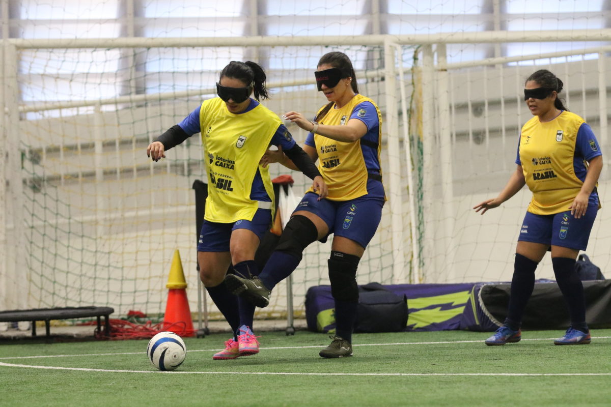Foto colorida de três atletas da seleção brasileira de futebol de cegas disputando a bola em treinamento em quadra de São Paulo. Elas usam vendas nos olhos e um colete amarelo e azul. Uma delas está com a perna levantada e a bola está na sua frente