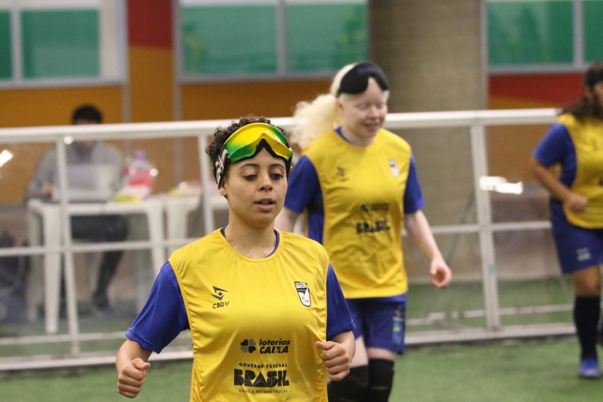 Foto colorida de atletas da seleção brasileira de futebol de cegas. Em primeiro plano, aparece Sarah Santana com a venda levantada e correndo. Ela é branca e tem cabelo preto curto. Ao fundo outra atleta com a venda levantada. Elas usam camisetas amarelas com detalhes azuis
