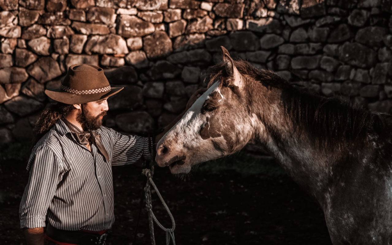 Foto colorida de homem acariciando a cabeça de um cavalo. O homem está vestido ao estilo gaúcho, com chapéu e uma camisa branca. 