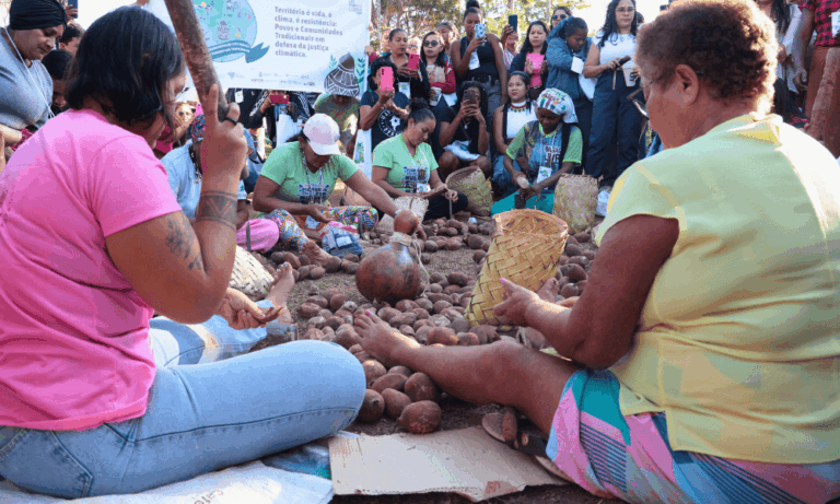 Foto colorida de quebradeiras de coco babaçu do MIQCB durante evento em Brasília (DF). Na imagem, as quebradeiras aparecem sentadas no chão e quebrando cocos, enquanto pessoas assistem.