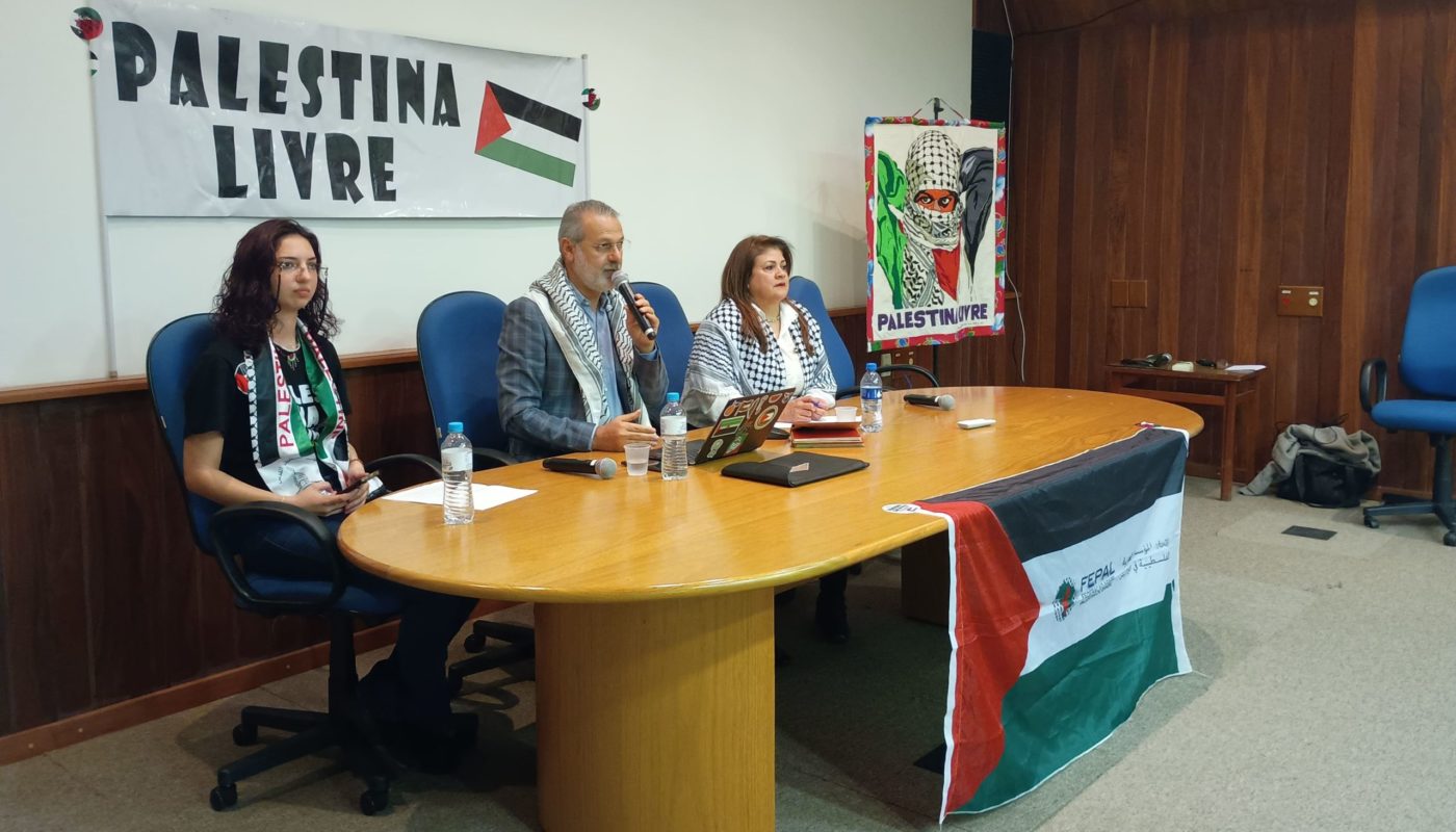 Foto colorida de Ualid Rabah em mesa durante debate na Universidade Federal de Santa Maria. Ele é um homem branco, com cabelo e barba grisalha. Ao seu lado, estão a vereadora Helen Cabral e a representante do Comitê Palestina Livre de Santa Maria. Na imagem também aparecem a bandeira da Palestina e um cartaz escrito Palestina Livre