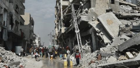 Foto colorida de pessoas buscando água ao lado de um prédio que corre o risco de desabar em Beit Lahia, no Norte de Gaza.