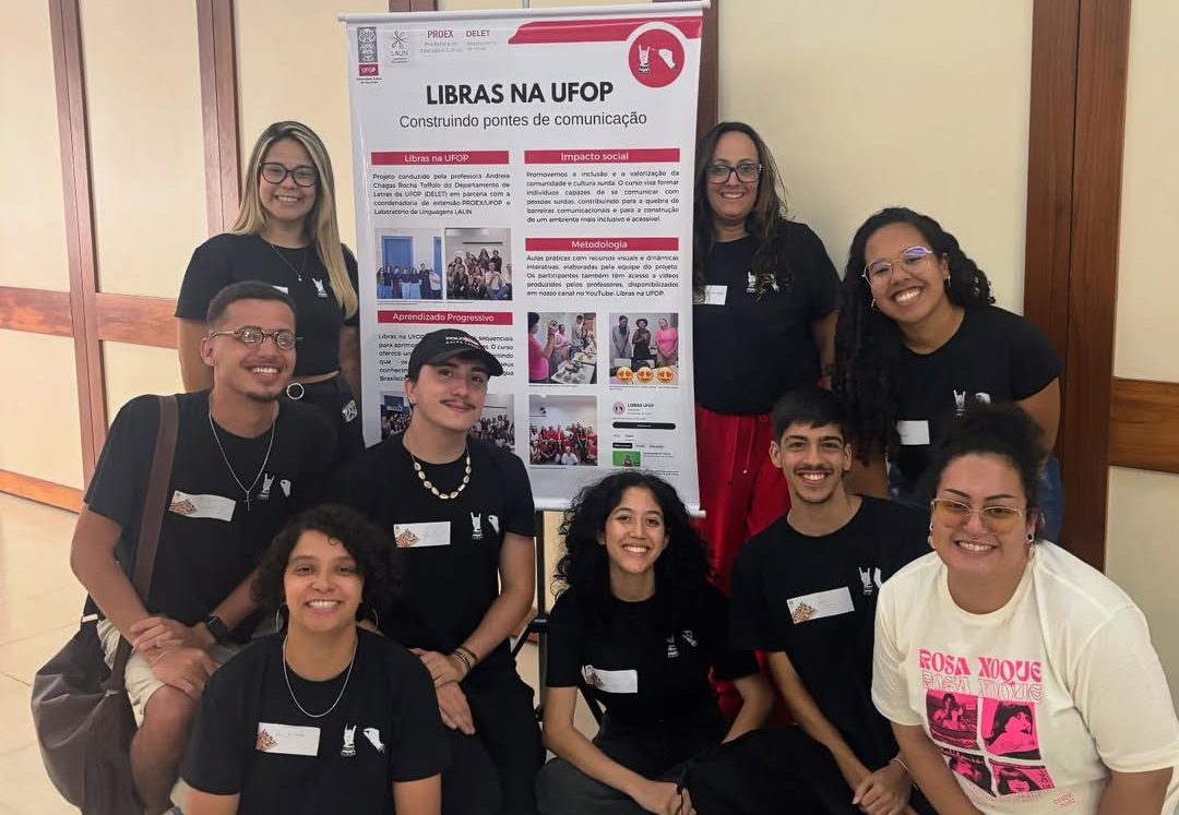 Foto colorida de membros do projeto de extensão Libras na UFOP. Na imagem, diversos estudantes aparecem posando para a foto ao lado de um cartaz com o nome do projeto