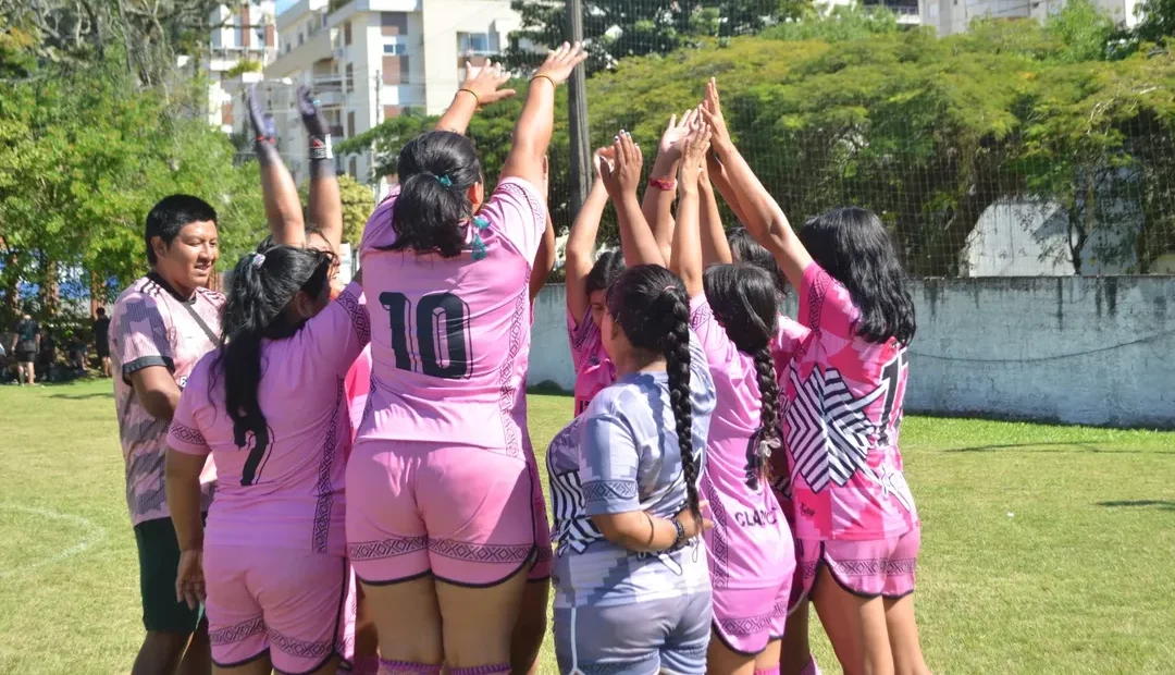 Foto colorida das atletas da equipe Kunhangue Itaty. As mulheres guaranis da equipe aparecem de costas e reunidas, com as mãos para o alto. Ela usam uniforme rosa e se reunem para jogo da Liga Mbya Guarani