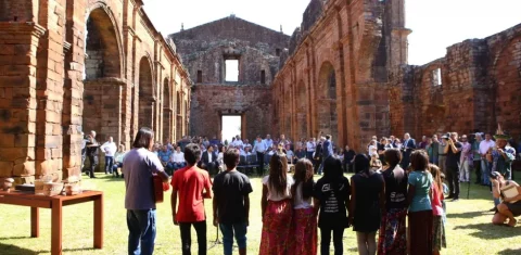 Foto colorida do Coral Mbyá-Guarani durante cerimônia de reconhecimento do Sítio Arqueológico de São Miguel das Missões como patrimônio do Mercosul. Na imagem, os guaranis aparecem de costas no interior das ruínas da Igreja e ao fundo, aparecem diversas pessoas.