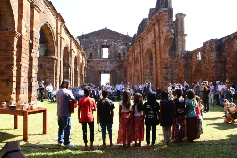 Foto colorida do Coral Mbyá-Guarani durante cerimônia de reconhecimento do Sítio Arqueológico de São Miguel das Missões como patrimônio do Mercosul. Na imagem, os guaranis aparecem de costas no interior das ruínas da Igreja e ao fundo, aparecem diversas pessoas.