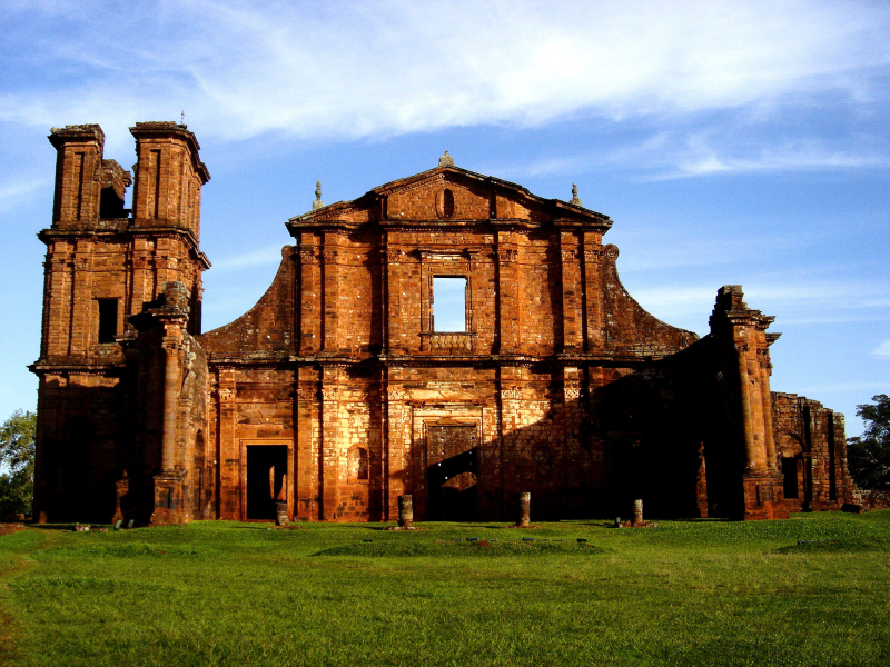 Foto colorida das ruinas de igreja missioneira em São Miguel das Missões (RS). A imagem mostra as ruínas da frente da Igreja, com estilo barroco. Ao fundo, céu azul