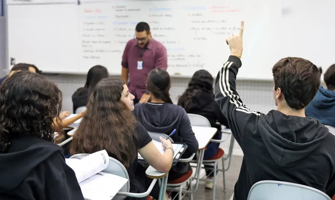 Foto colorida de sala de aula. Um menino está com a mão levantada e olha para o professor que aparece na frente da turma desfocado. Outros estudantes estão na sala e no fundo aparece o quadro