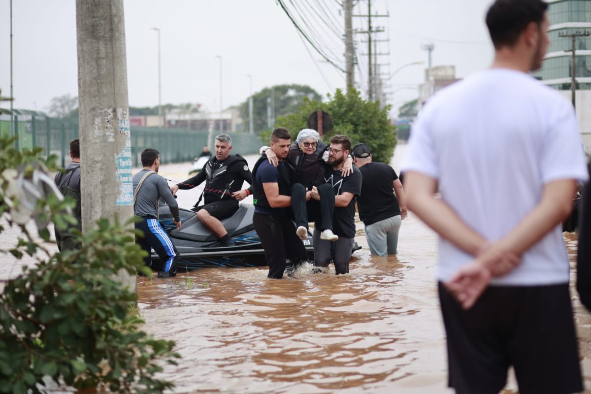 Foto colorida do resgate de uma pessoa idosa durante as enchentes no RS. Na imagem, dois homens carregam a mulher nos braços e outros aparecem em uma lancha atrás, o chão está coberto de água. Iniciativa propõe protocolos para resgate inclusivo.