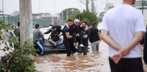 Foto colorida do resgate de uma pessoa idosa durante as enchentes no RS. Na imagem, dois homens carregam a mulher nos braços e outros aparecem em uma lancha atrás, o chão está coberto de água. Iniciativa propõe protocolos para resgate inclusivo.