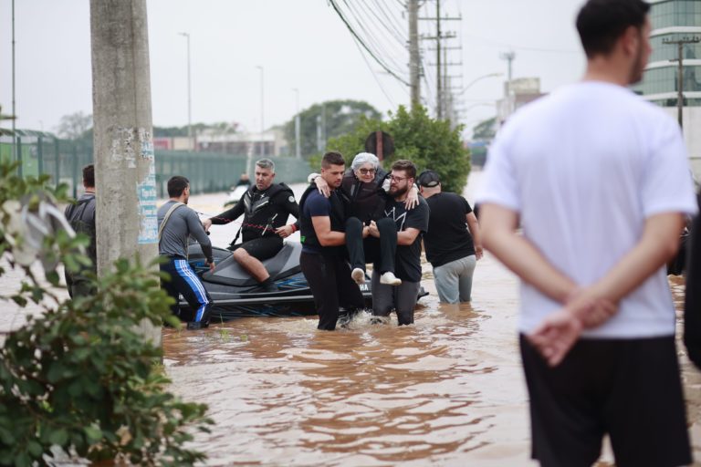 Foto colorida do resgate de uma pessoa idosa durante as enchentes no RS. Na imagem, dois homens carregam a mulher nos braços e outros aparecem em uma lancha atrás, o chão está coberto de água. Iniciativa propõe protocolos para resgate inclusivo.