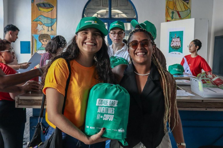 Foto colorida de duas mulheres na Cidade das Juventudes. Uma delas, jovem e branca, segura uma mochila e usa um boné que diz "Cidade das Juventudes". Ao lado, aparece uma mulher negra de óculos escuros. Ao fundo, outros jovens