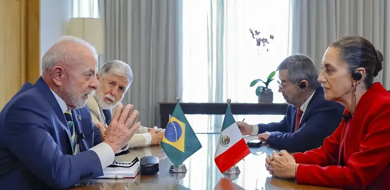 Foto colorida do presidente Lula e da presidenta do México, Claudia Sheinbaum sentados em uma mesa de frente um para o outro. Ambos conversas e as bandeiras de seus países estão diante deles