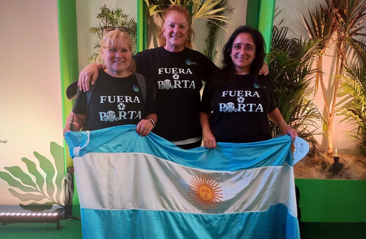Foto colorida de Silvia Cruz, Maria Rosa Vignolo e Natalia Tello. Elas usam uma camiseta preta, escrito em branco "Fuera Porta" e seguram a bandeira da argentina