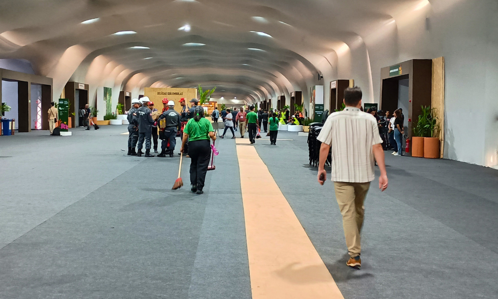 Foto colorida com pessoas de costas no corredor da zona azul da COP30. No centro da imagem, aparece uma trabalhadora da limpeza