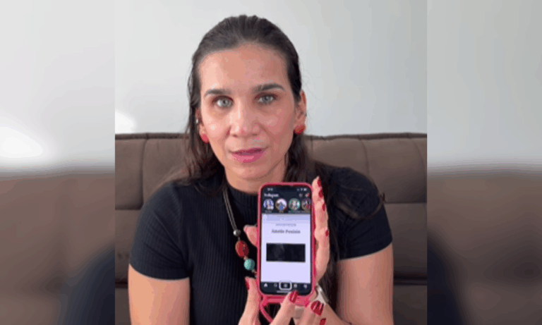 Captura de tela colorida com foto de Geisa Farini. Ela é uma mulher branca com cabelos pretos. Ela está segurando um celular com o Instagram aberto e com a tela voltada para frente. Ela usa um colar com contas coloridas e uma camiseta preta. Ao fundo, sofá marrom e parede branca