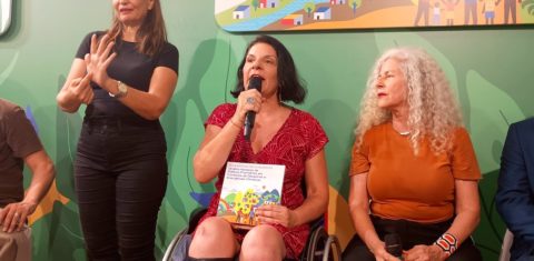 Foto colorida de Anna Paula Feminella ao lado de Scheilla Abbud, sentadas, e da intérprete de Libras durante painel sobre emergência climática e resgate de pessoas com deficiência. Ana Paula é uma mulher branca com cabelos curtos pretos, usa um vestido vermelho e cadeira de rodas. Ela segura o microfone com uma mão e um livro com outra. Sheila é uma mulher branca, com cabelos loiros cacheados, usa uma camiseta laranja e calça preta. A intérprete de Libras está toda de preto e sinalizando em pé ao lado de Ana Paula. Acessibilidade na COP30