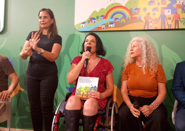 Foto colorida de Anna Paula Feminella ao lado de Scheilla Abbud, sentadas, e da intérprete de Libras durante painel sobre emergência climática e resgate de pessoas com deficiência. Ana Paula é uma mulher branca com cabelos curtos pretos, usa um vestido vermelho e cadeira de rodas. Ela segura o microfone com uma mão e um livro com outra. Sheila é uma mulher branca, com cabelos loiros cacheados, usa uma camiseta laranja e calça preta. A intérprete de Libras está toda de preto e sinalizando em pé ao lado de Ana Paula. Acessibilidade na COP30