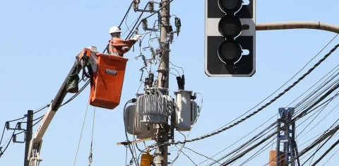 Foto colorida de trabalhador do setor elétrico em cesto, trabalhando em linha de rede elétrica, ao fundo, céu azul
