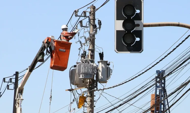 Foto colorida de trabalhador do setor elétrico em cesto, trabalhando em linha de rede elétrica, ao fundo, céu azul