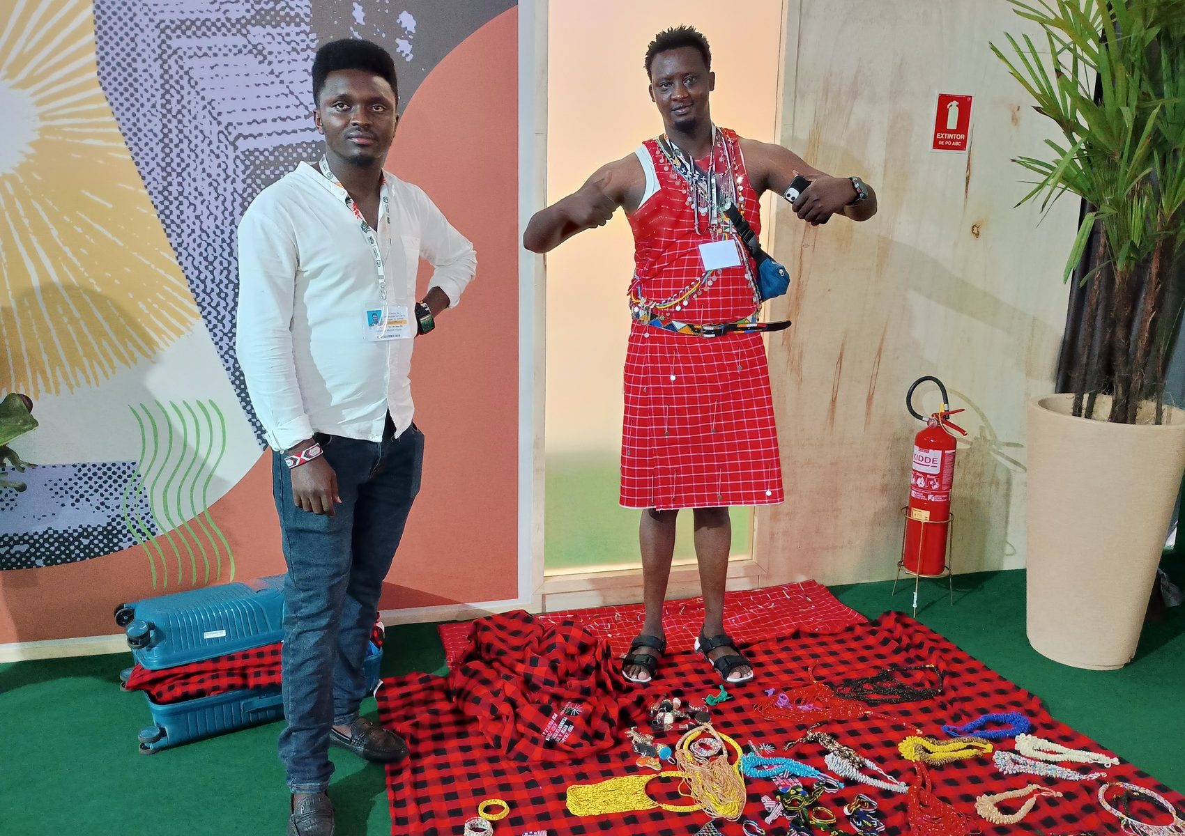 Foto colorida de Abraham Maison Kipila ao lado de outro ativista queniano. Ele é um homem preto de cabelo preto curto, usa uma camissa branca e calça jeans azul escura. Ao seu lado, o outro ativista está com uma veste tradicional Maasai, vermelha.