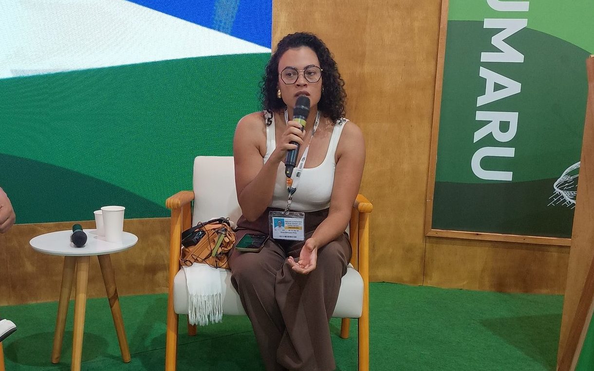 Foto colorida de Vânia Marques Pinto. Ela é uma mulher negra, com cabelo preto cacheado na altura do ombro. Ela usa uma blusa branca e calça marrom, está sentada e segura um microfone