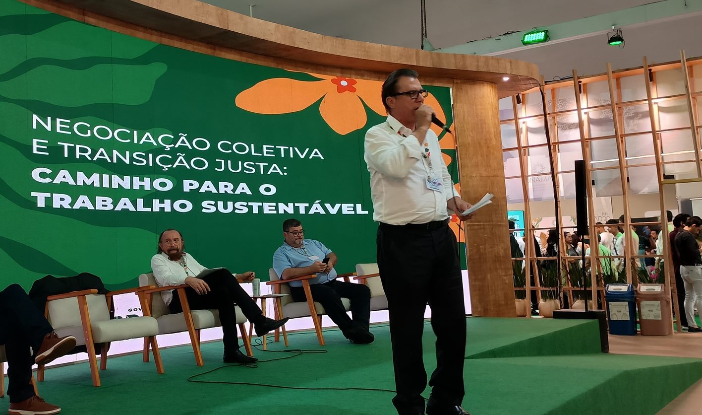 Foto colorida com o ministro Luiz Marinho em pé. Ele é um homem branco, com cabelo curto preto, usa camisa branca e calça preta. Ao fundo, painel sobre transição justa e outros dois homens sentados