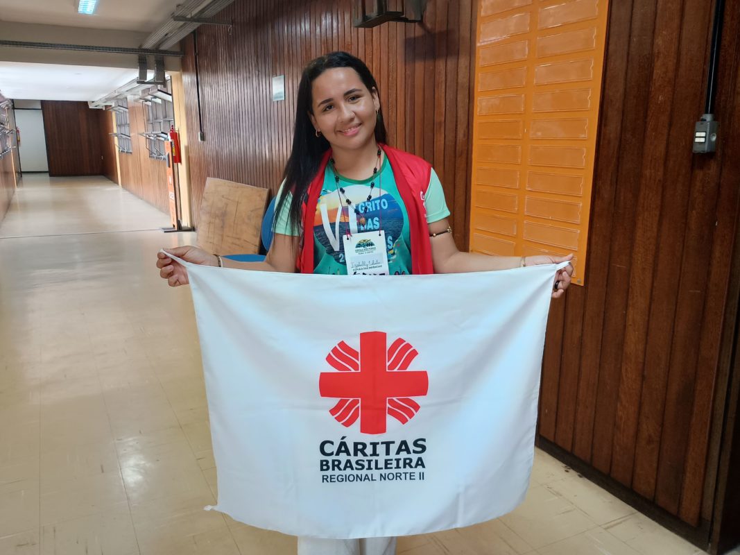 Foto colorida de Izabelly Lobato dos Santos. Ela é uma jovem ribeirinha de cabelos pretos e segura uma bandeira branca com a logo vermelha da Cáritas Vermelhas
