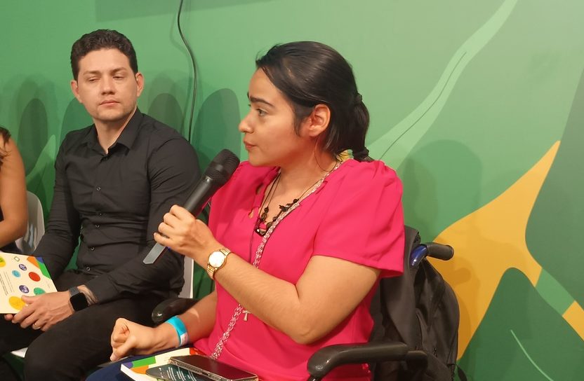 Foto colorida de Jéssica Ribeira. Ela é uma mulher branca com cabelo preto e usuária de cadeiras de rodas. Ela aparece de lado e segurando um microfone. Ela usa uma camiseta rosa. Ao seu lado, aparece um homem branco de camiseta preta. Fundo verde.