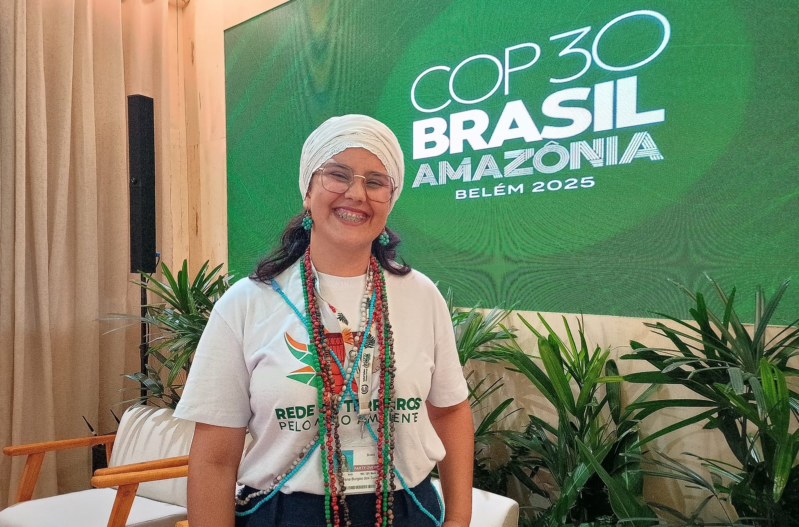 Foto colorida de Rayana Burgos. Ela é uma mulher negra, com cabelo preto. Ela está usando um pano branco no cabelo, uma camiseta branca e colares coloridos de povos de terreiro. Ao fundo, logo da COP30