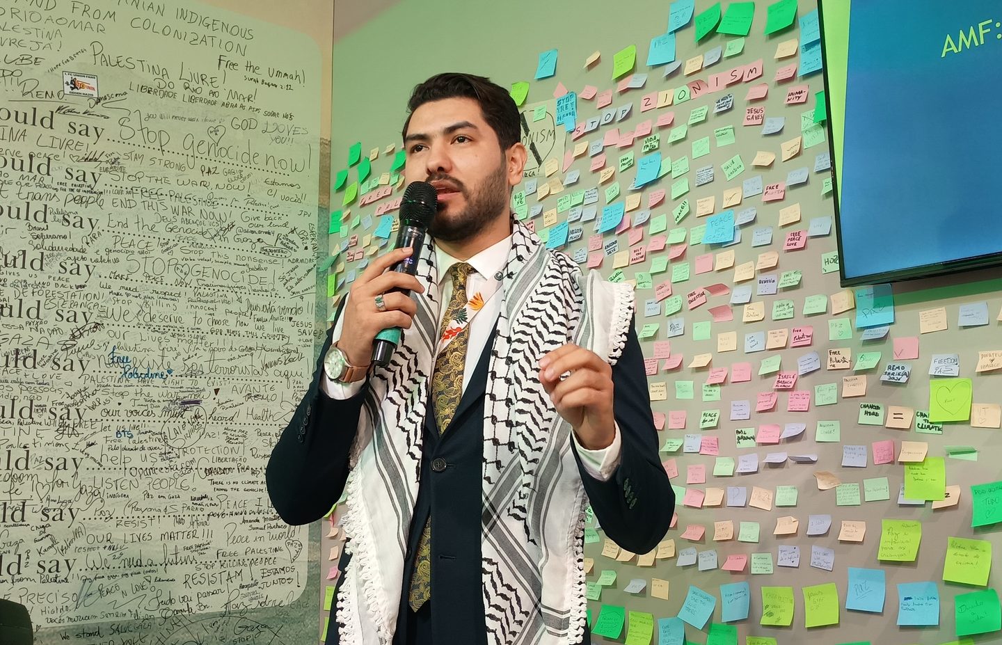 Foto colorida de Tareg Mustafa. Ele é um homem branco, com cabelo e barba preta. Ele está usando um terno azul escuro e um lenço preto e branco, enquanto segura o microfone