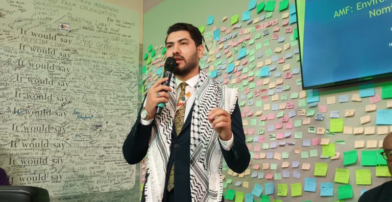 Foto colorida de Tareg Mustafa. Ele é um homem branco, com cabelo e barba preta. Ele está usando um terno azul escuro e um lenço preto e branco, enquanto segura o microfone