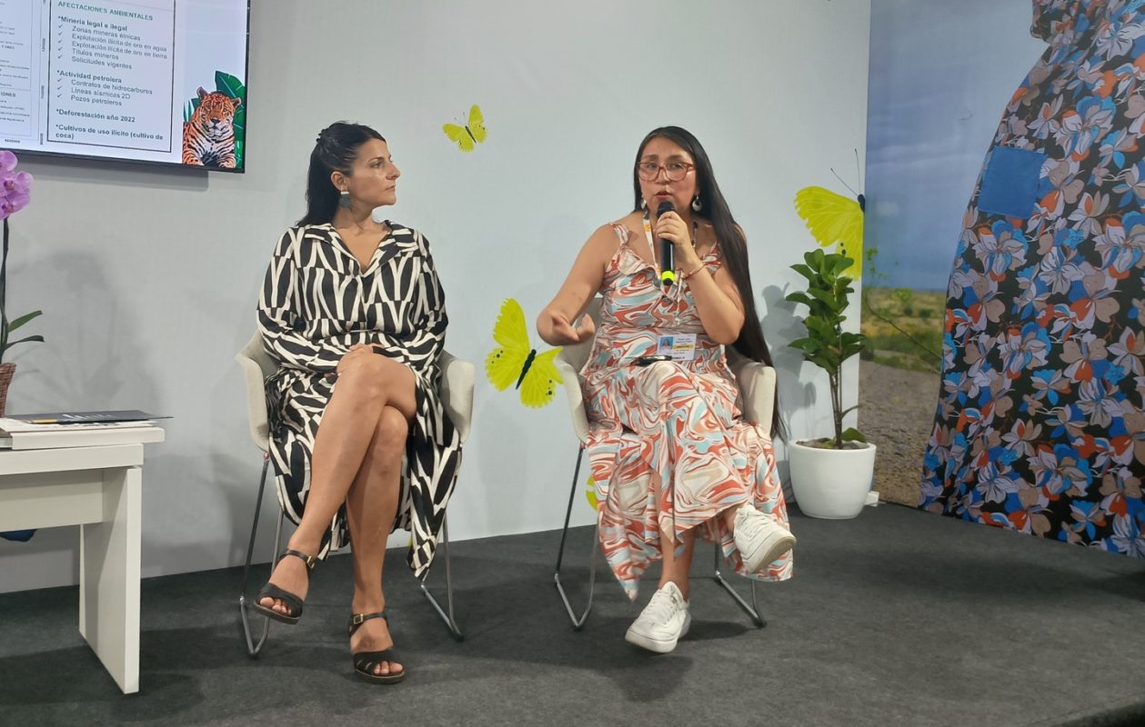 Foto colorida de Ruth Luque, com vestido branco e laranja e segurando o microfone, ao lado da ministra Irene Vélez. Ambas são mulheres brancas de cabelos pretos