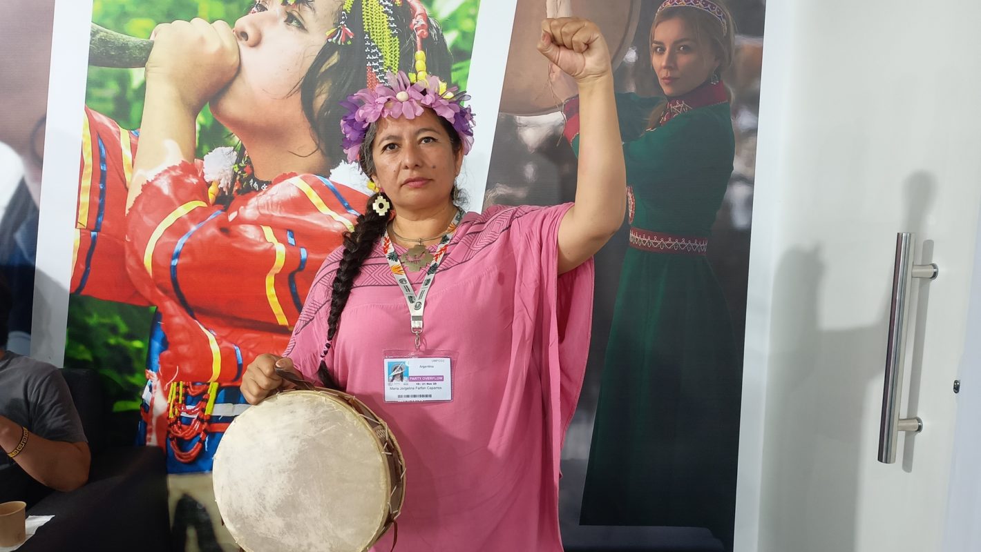 Foto colorida de Maryta de Humahuaca. Ela é uma indígena de cabelo preto enrolado em uma trança. Ela usa um vestido rosa e uma tiara com penas rosas. Ela olha para frente e ergue o braço direito.