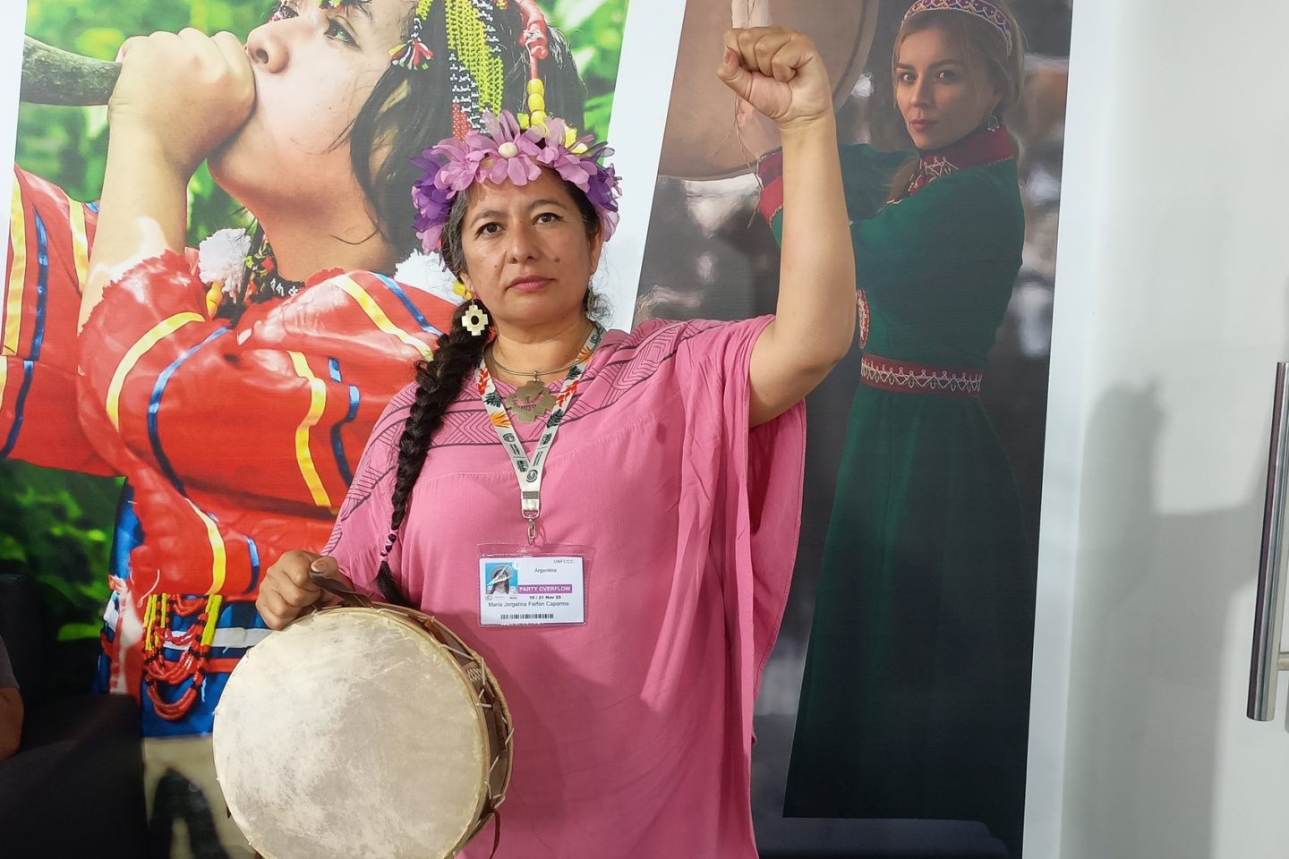 Foto colorida de Maryta de Humahuaca. Ela é uma indígena de cabelo preto enrolado em uma trança. Ela usa um vestido rosa e uma tiara com penas rosas. Ela olha para frente e ergue o braço direito.