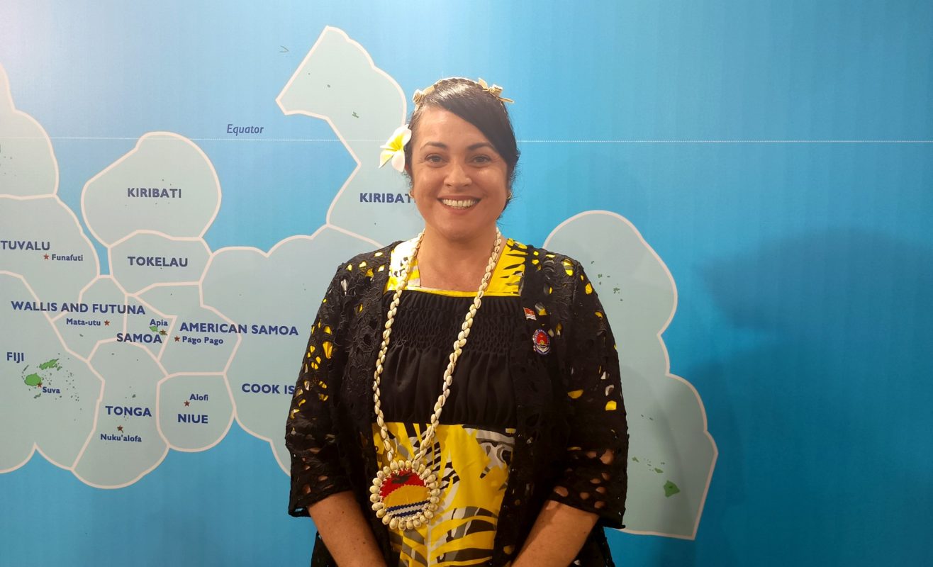 Foto colorida de Ruth Cross Kwansing. Ela é uma mulher branca de cabelos pretos que estão atados. Ela sorri e usa um vestido preto, com detalhes em amarelo, e um colar com a bandeira do Kiribati. Ao fundo, mapa das ilhas do Pacífico